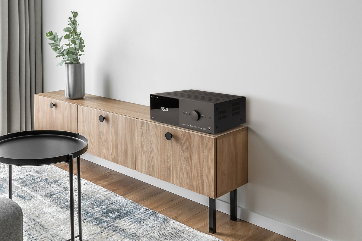 A modern wooden media console with black legs and knobs, styled in a minimalist living room setting with a black Anthem AV processor on top, a potted plant, and a small black side table.