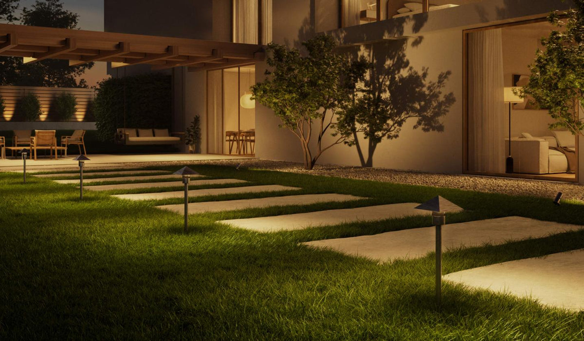Modern outdoor landscape at dusk showing a landscaped lawn with illuminated stone stepping stones, small ground lights, trees, and a contemporary house with warm interior lighting.