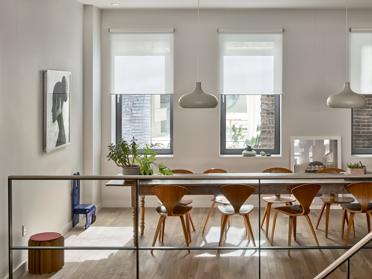 A modern dining room interior with a long wooden dining table and multiple mid-century style wooden chairs, set under pendant lights with large windows and Lutron roller shades in the background.