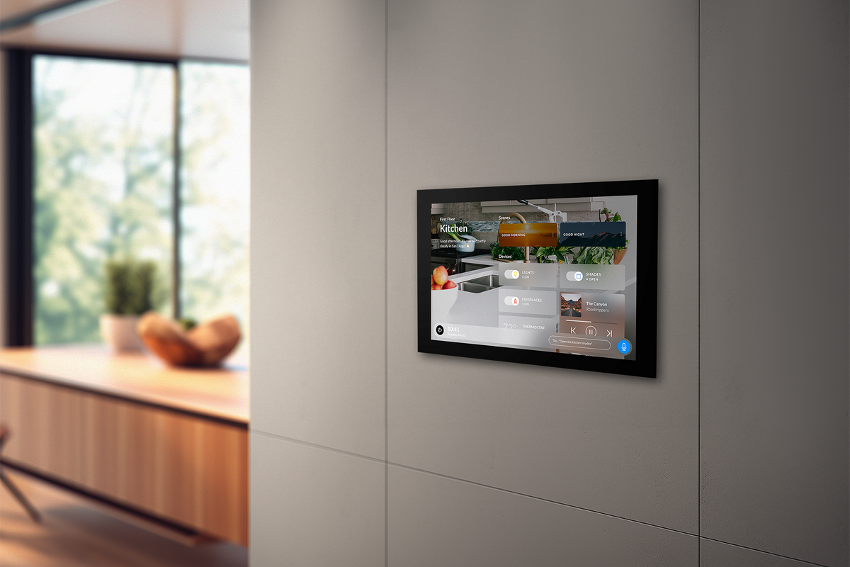 A black-framed Josh.ai touchscreen mounted on a light gray paneled wall, showing a kitchen scene on its screen. The background is a modern kitchen interior with a wooden counter and large windows.