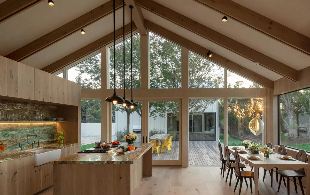 A modern open-plan kitchen and dining area with a high vaulted ceiling, large floor-to-ceiling windows, and natural wood cabinetry and flooring. Pendant lights hang over the kitchen island, and a dining table with chairs sits near the windows overlooking a deck and trees.