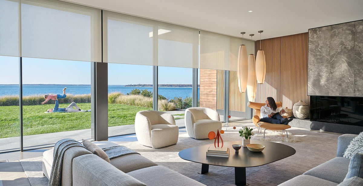 A modern living room with large roller blinds overlooking a grassy lawn and the ocean with husband and kids playing in te background. A woman is seated in a lounge chair near a fireplace, and the room features contemporary furniture including a sofa, armchairs, and a round coffee table.