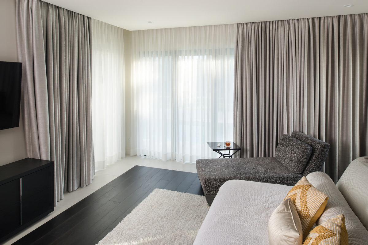 A modern living room featuring floor-length taupe curtains with matching sheer white curtains, a gray textured armchair, a light-colored sofa with patterned pillows, and dark wood flooring.
