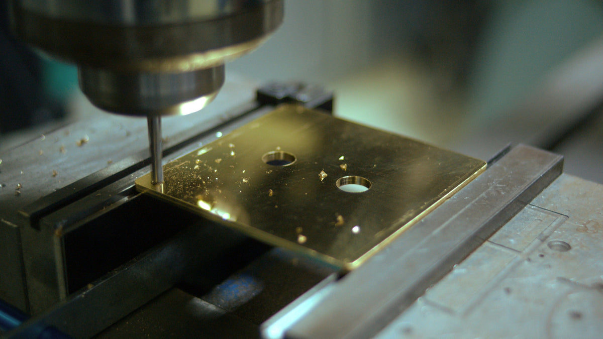 A close-up of a CNC milling machine's drill bit actively drilling a hole into a polished brass Lutron switch plate held in a machine vise.
