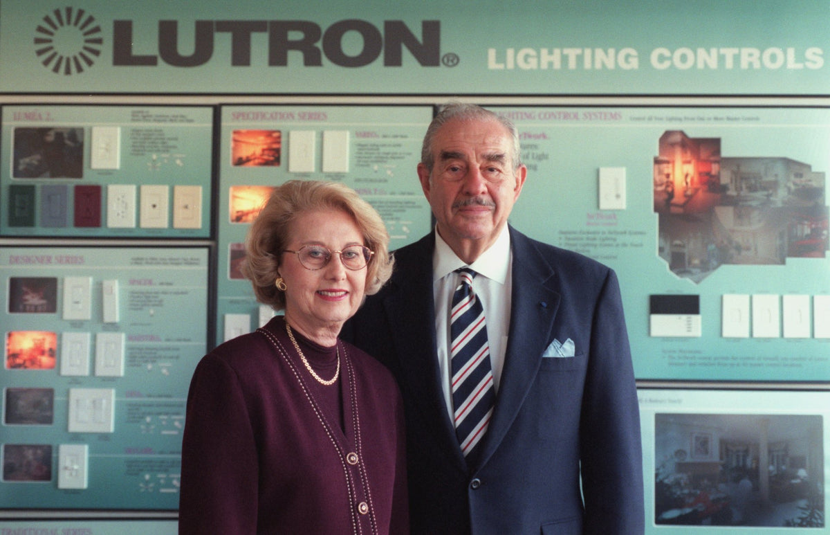 Vintage photo of Ruth and Joel Spira standing in front of a large Lutron Lighting Controls display wall. Joel is wearing a dark navy suit with a striped tie, and Ruth is wearing a dark plum button-up top with a gold necklace.