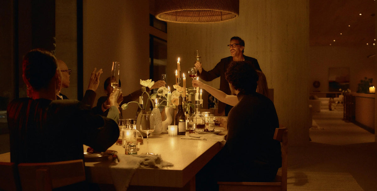 A warm, dimly lit dinner scene with several people gathered around a table set with candles, glasses, and floral arrangements. One person stands at the head of the table holding a glass, while others sit and raise glasses in a toast.
