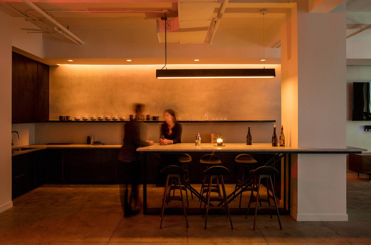 A modern kitchen interior with a long island, bar stools, dark cabinetry, warm under-cabinet Lutron lighting, and two blurred people standing near the counter.