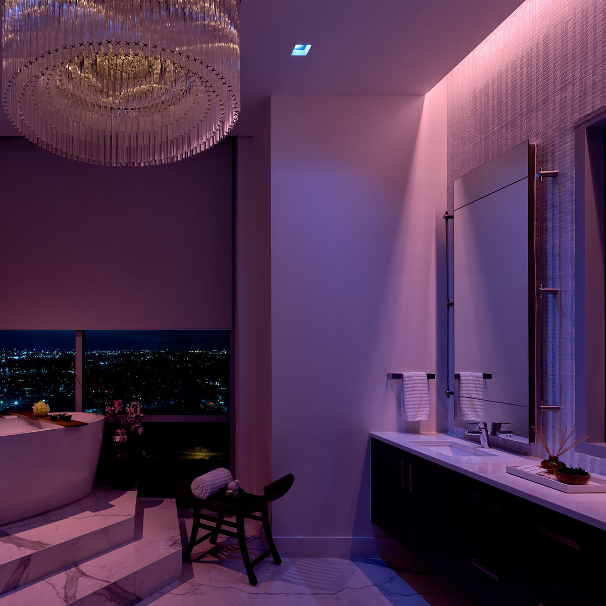 A luxury modern bathroom interior with a large crystal chandelier, a floating double-sink vanity, a large wall mirror, and a bathtub overlooking a city skyline at night.