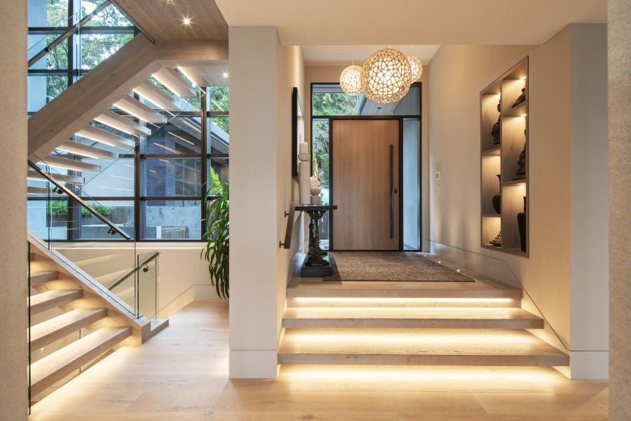 Modern home entryway with illuminated stone steps, glass railing staircase, warm wood flooring, and a round woven pendant light hanging above the front door.
