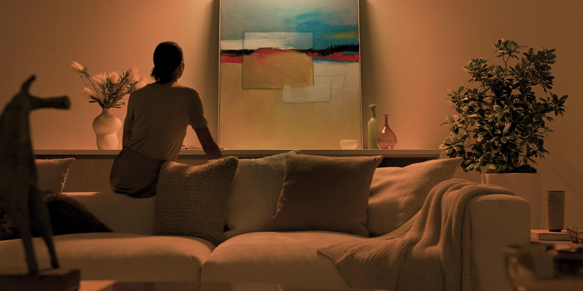 A woman sits on a beige sofa in a warmly lit living room, facing a large abstract painting on the wall. The scene includes pillows, a throw blanket, a plant, and decorative objects.