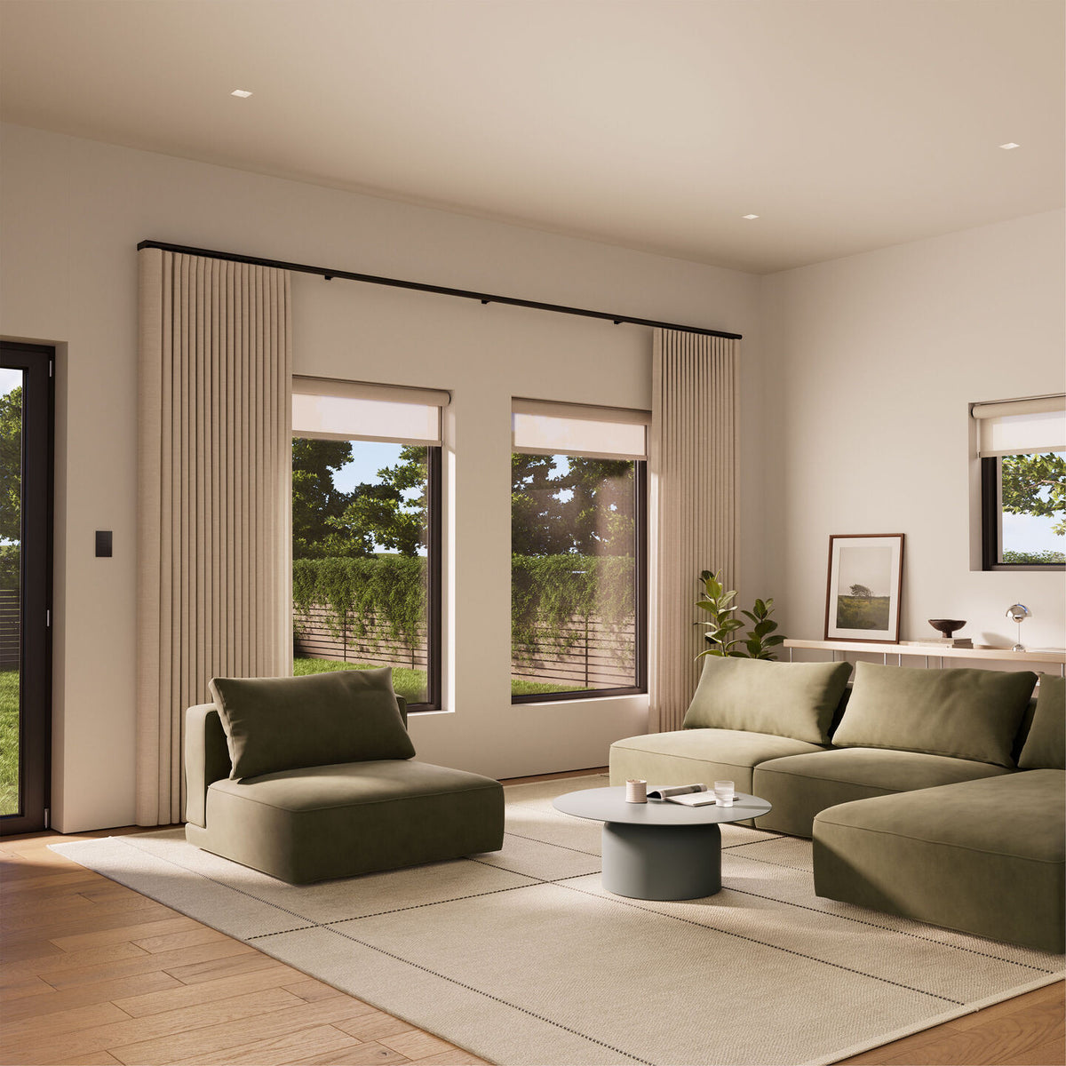 A modern living room with olive green sectional and lounge seating, a round coffee table, a large beige rug, and tall beige pleated Lutron Palladiom motorized drapes hanging in front of tall windows.