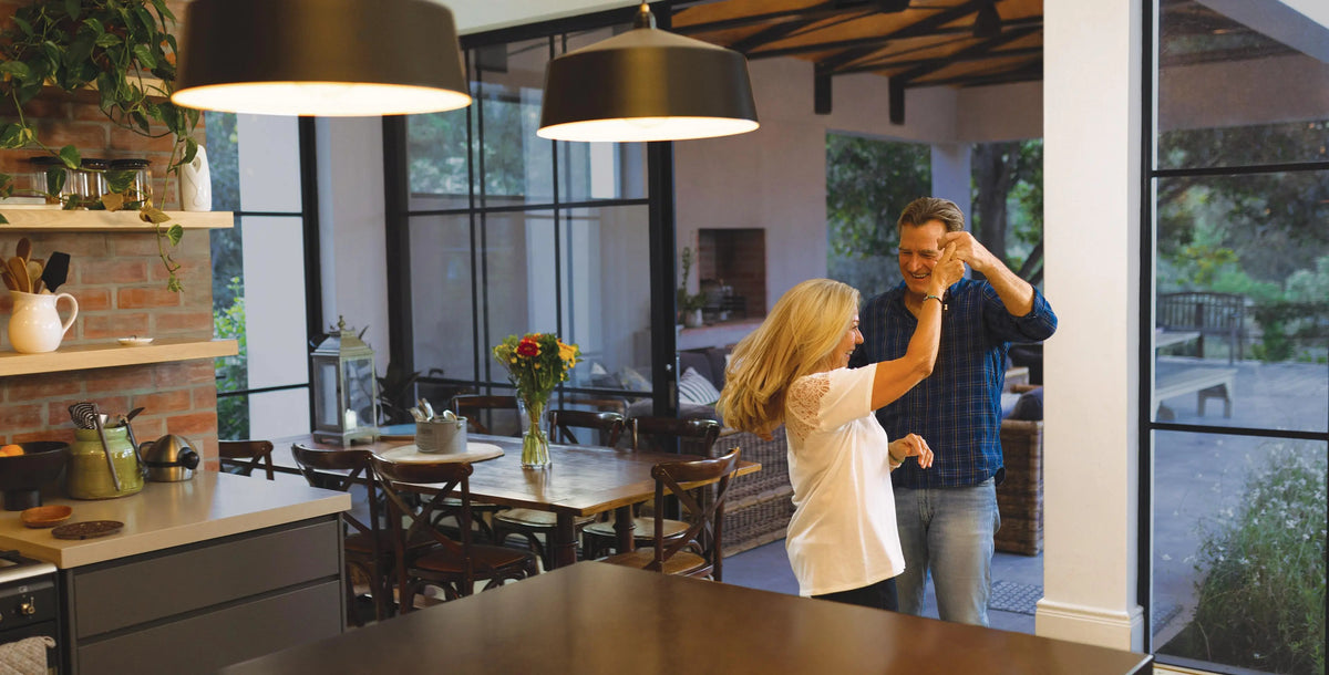 A man and a woman dancing together in a modern, bright kitchen and dining area with large windows and black pendant lights.
