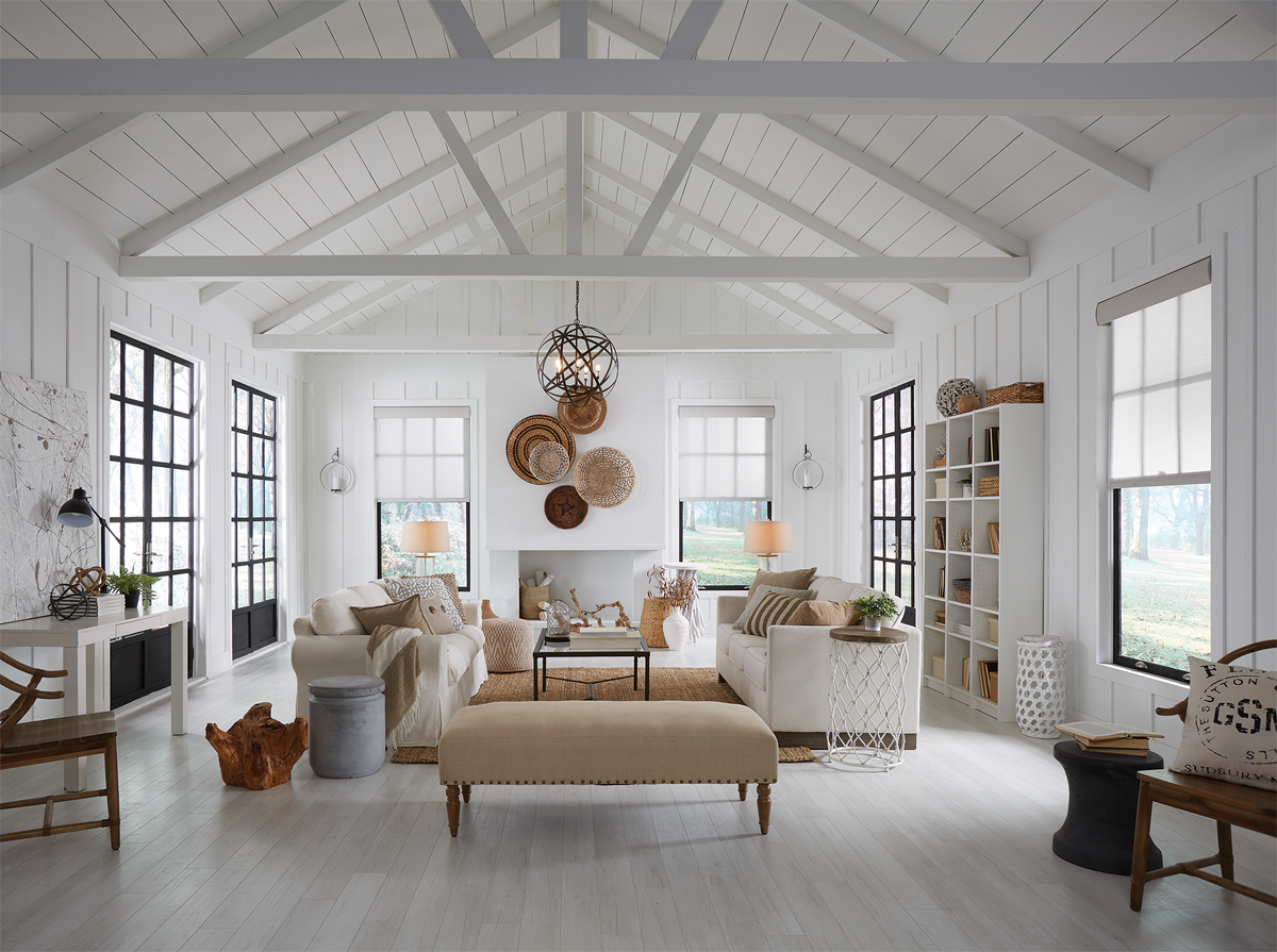 A bright, airy modern farmhouse-style living room with white paneled walls and ceiling, light wood floors, a cream upholstered bench, white sofas, black-framed windows, shelving, and decorative wall art.