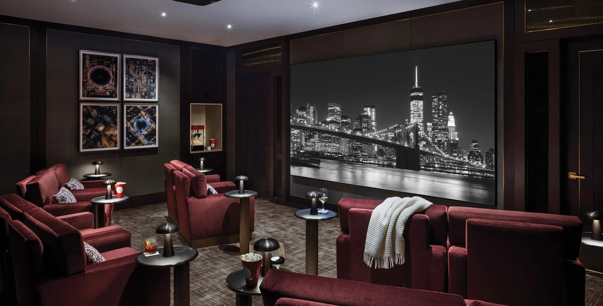 A luxury Dolby Atmos home theater interior with plush dark red velvet seating, round side tables, and a large black-and-white city skyline wall mural on the back wall and dramatic lighting throughout.
