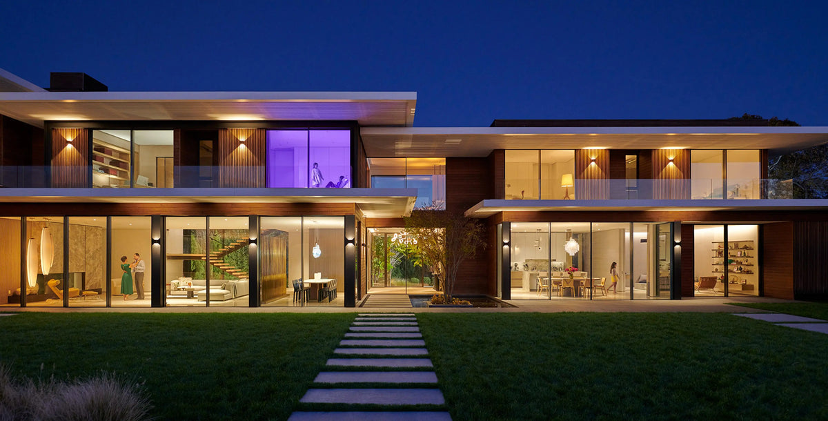 Modern two-story residential house at dusk with large floor-to-ceiling glass walls, warm interior lighting, a dark blue evening sky, and a landscaped lawn with a stone walkway in front.