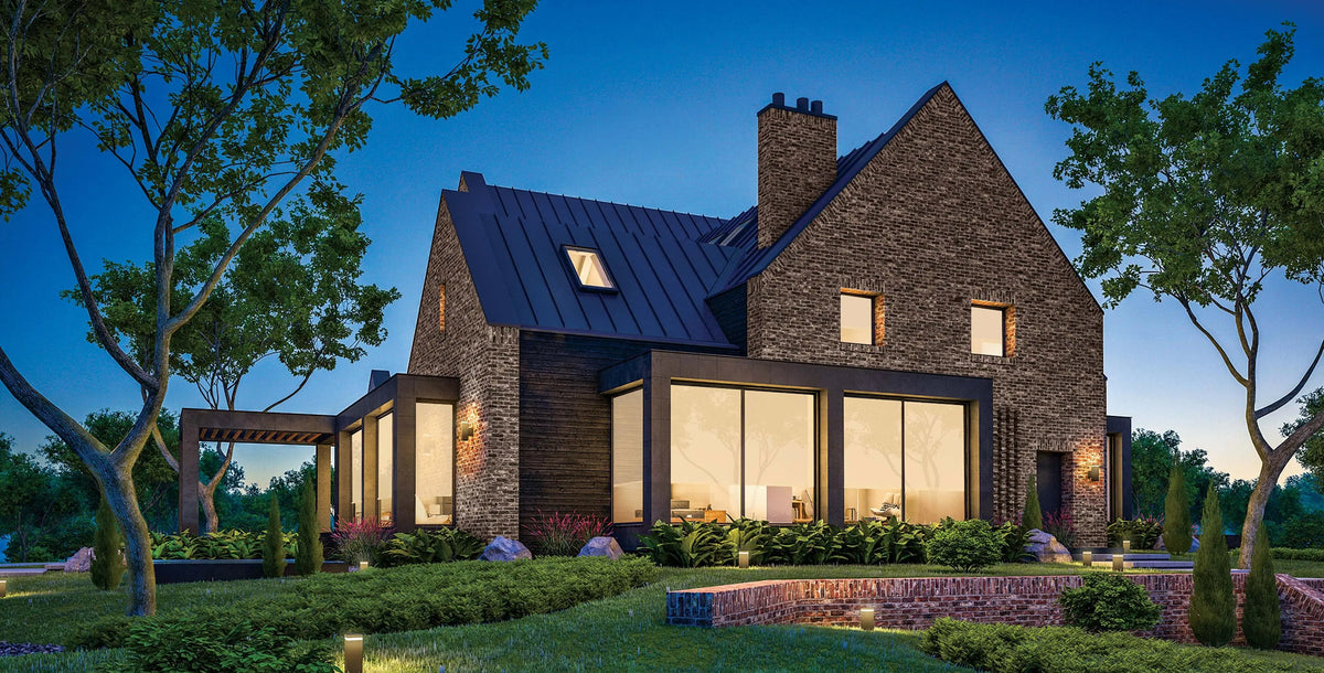 A modern two-story house with a dark metal roof, stone and wood exterior walls, large floor-to-ceiling windows, and a landscaped garden at dusk.