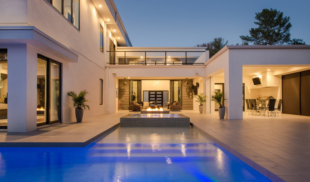 Modern luxury home exterior at dusk with a long blue-lit swimming pool in the foreground, white stucco architecture, glass sliding doors, potted plants, and an open patio area with dining furniture.