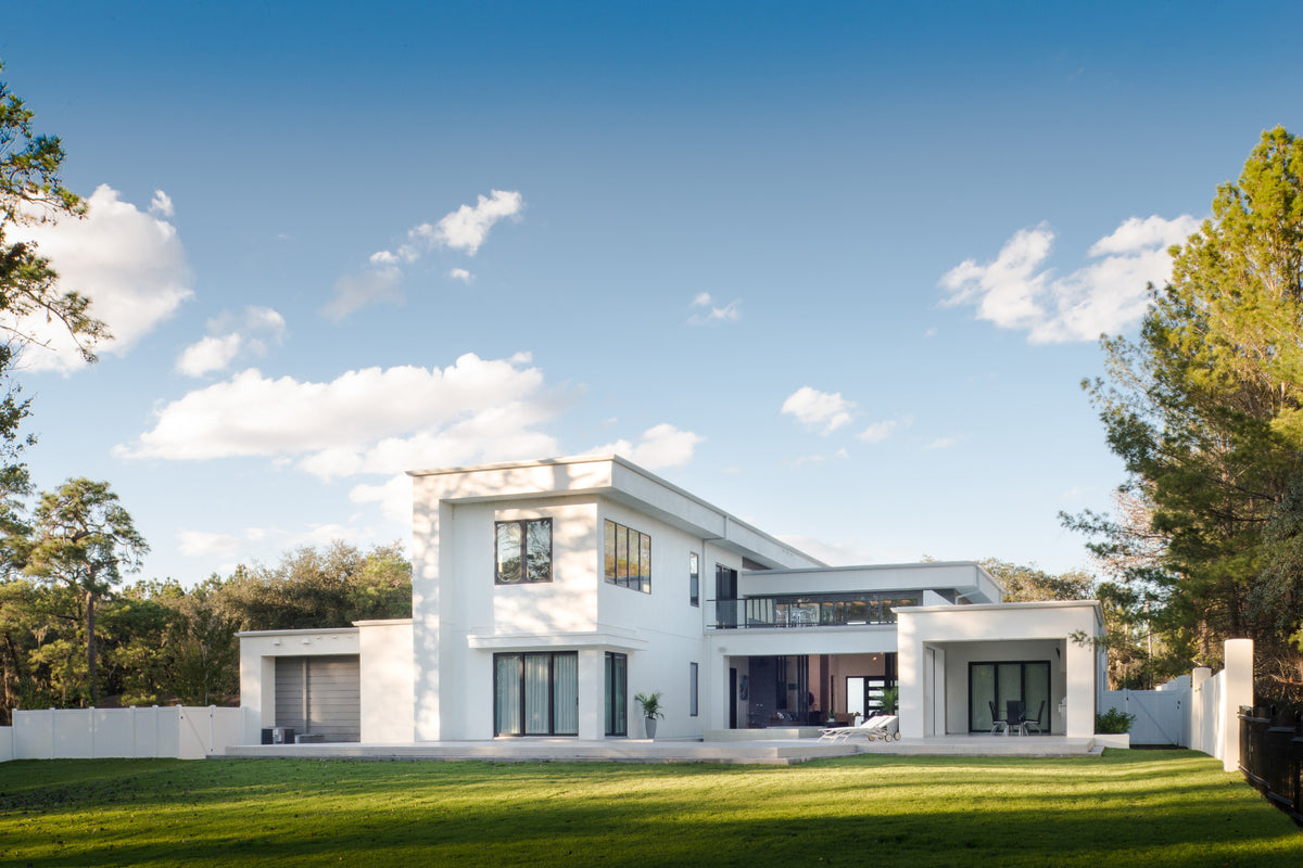 A modern two-story white house with large black-framed windows, set against a bright blue sky with scattered clouds and green trees and lawn in the foreground.