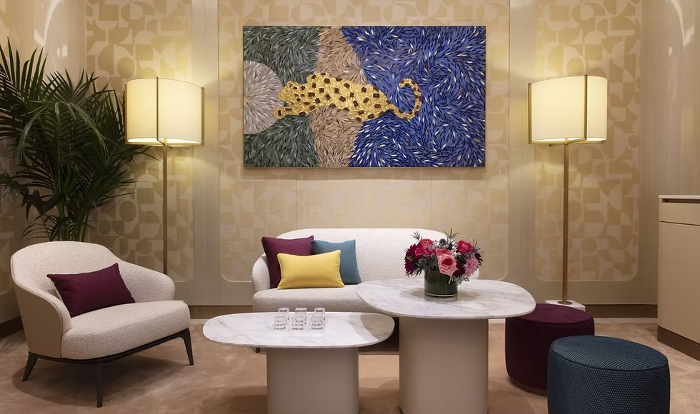 A modern living room interior featuring a large abstract canvas painting with blue, gold, and green brushstrokes hanging above a white sofa. The room includes a cream armchair with a burgundy pillow, two white oval coffee tables, a floral arrangement, two lit floor lamps, and a potted palm on a beige patterned wall.