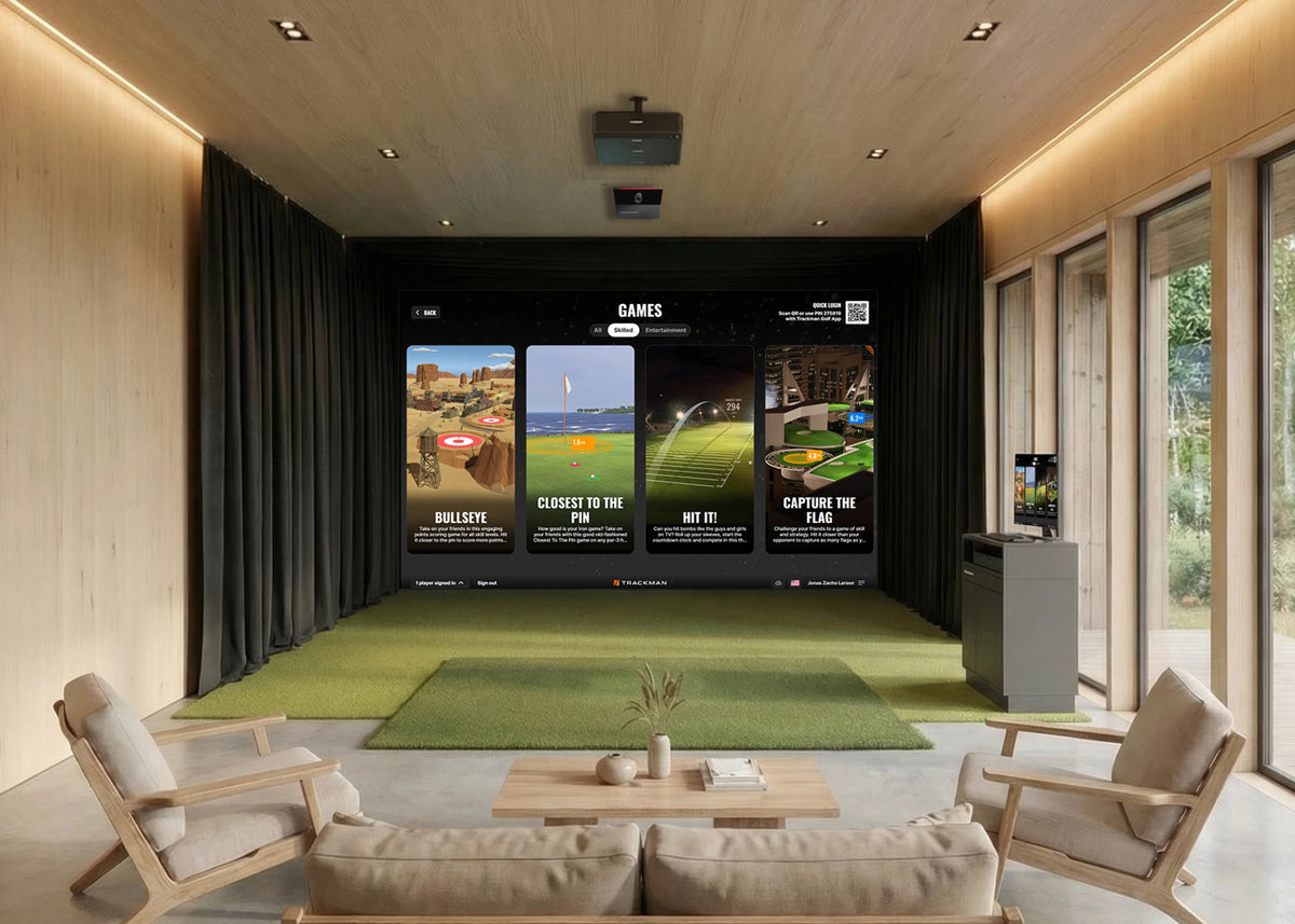 A modern indoor flex space home theater and simulation room with a large projection screen displaying golf game panels, green turf flooring, wooden ceiling and walls, black curtains, and two light wood lounge chairs facing the screen.