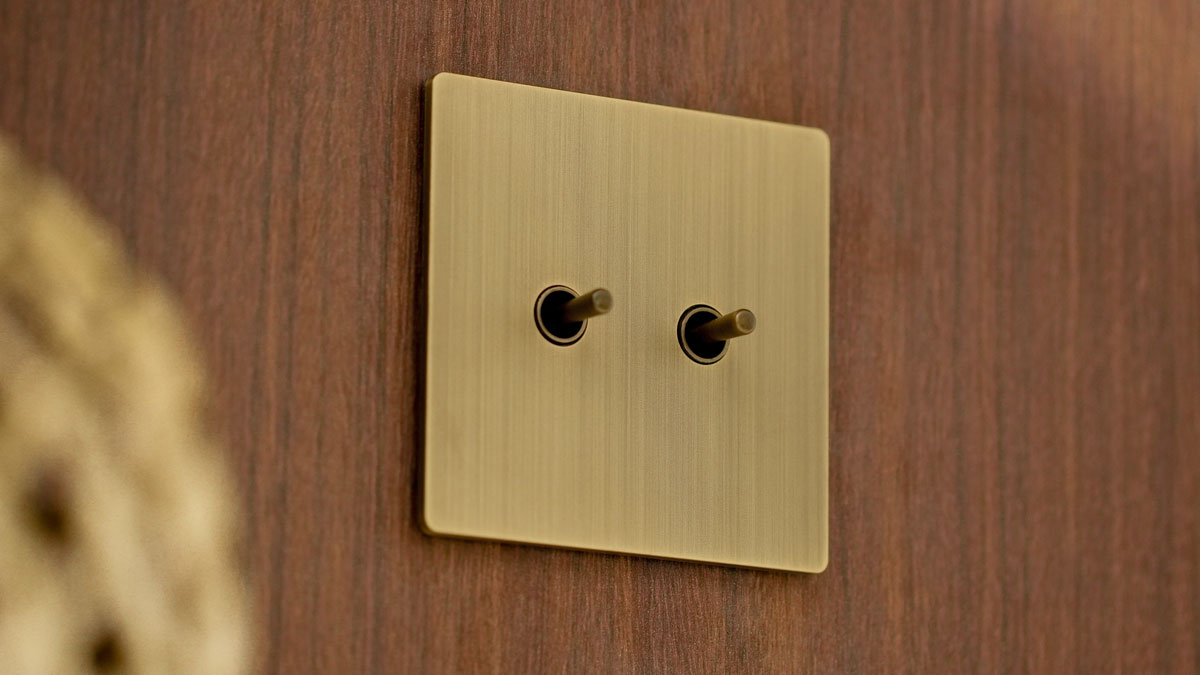A square brushed brass Lutron Aviena double-toggle switch wall plate mounted on a walnut wood panel.