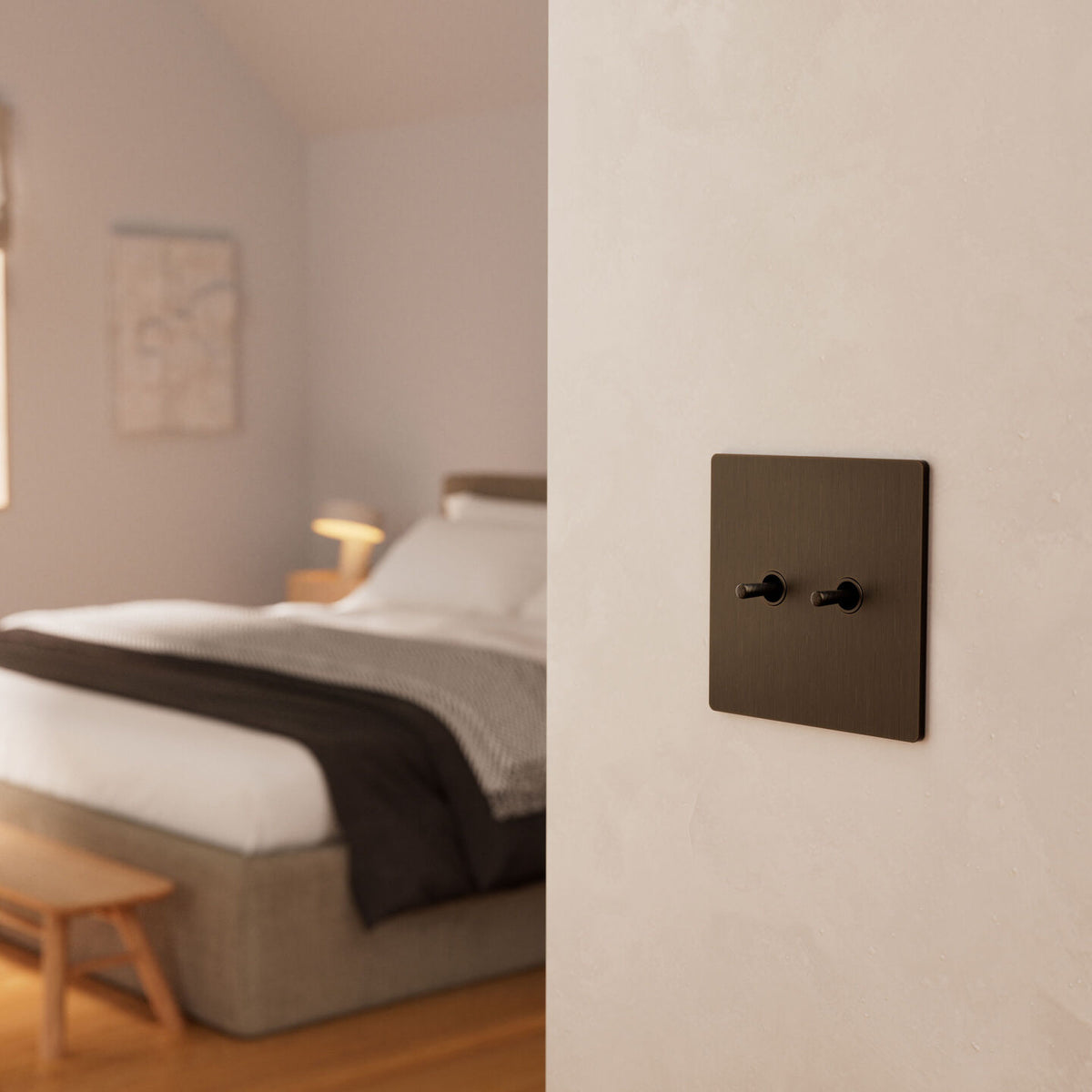 Aged Bronze Lutron Aviena toggle switches mounted on a textured off-white wall, with a softly blurred bedroom in the background.